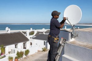 Technician on a ladder adjusting a satellite dish on a whitewashed guesthouse roof in a West Coast lagoon town with other dishes visible on nearby cottages