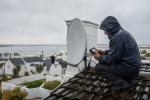 Satellite dish on a rooftop in a West Coast coastal town during rain, with a technician checking signal alignment